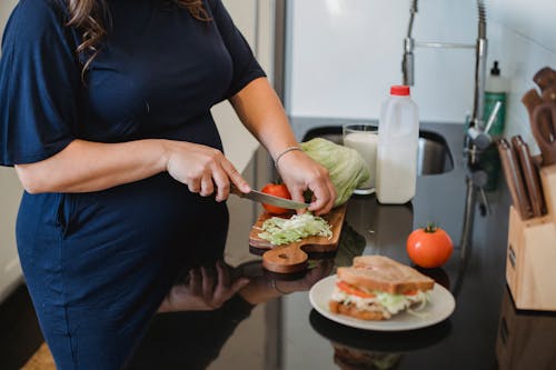 Free Crop anonymous pregnant female in blue dress standing near counter and chopping fresh lettuce leaves for sandwich in modern kitchen Stock Photo